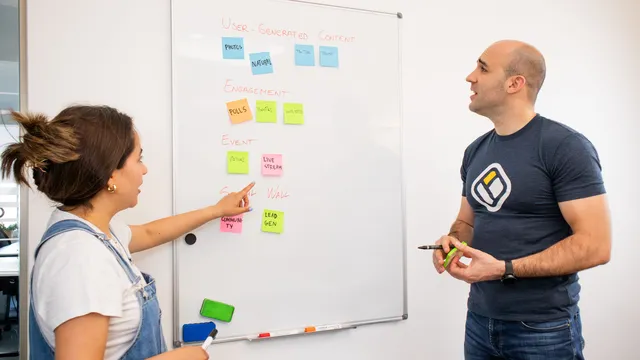 Zwei Kolleginnen und Kollegen besprechen an einem Whiteboard mit bunten Haftnotizen einen Plan