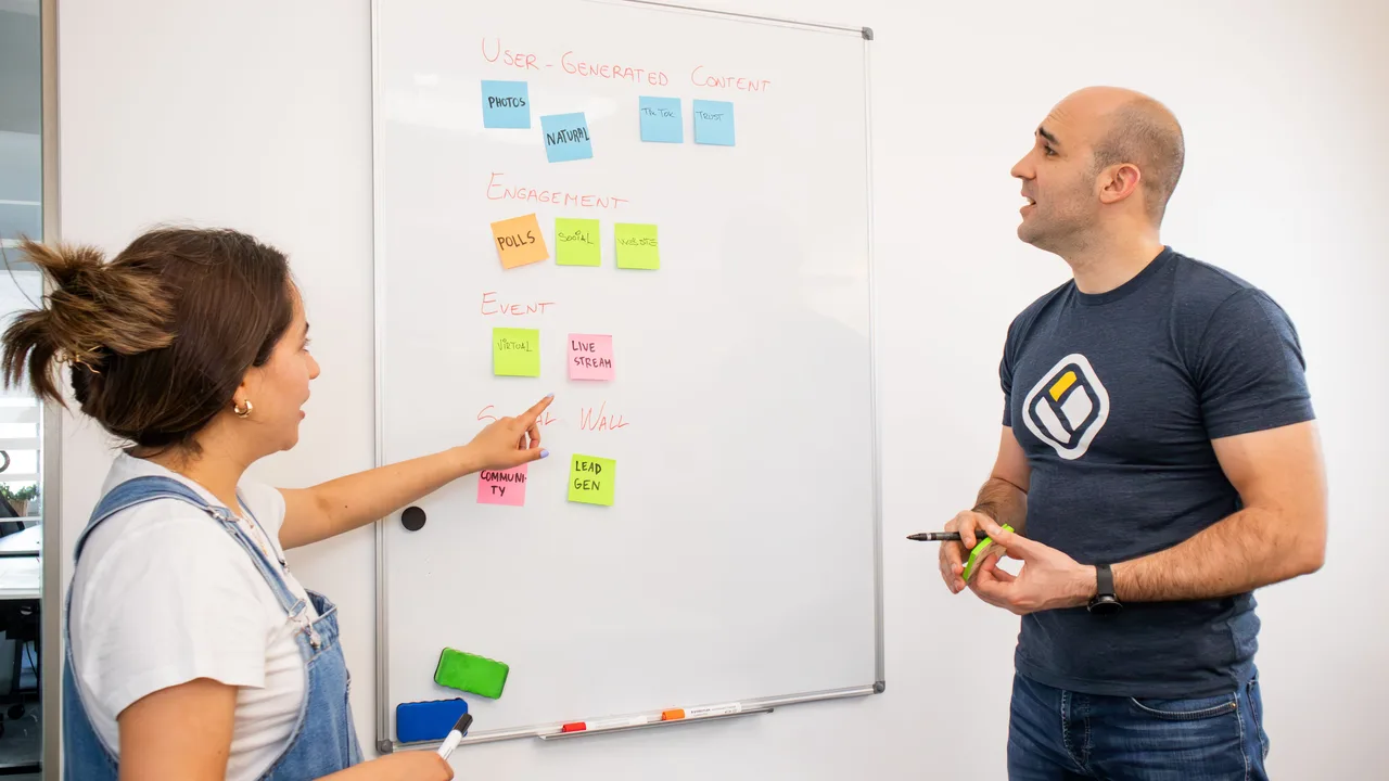 Zwei Kolleginnen und Kollegen besprechen an einem Whiteboard mit bunten Haftnotizen einen Plan