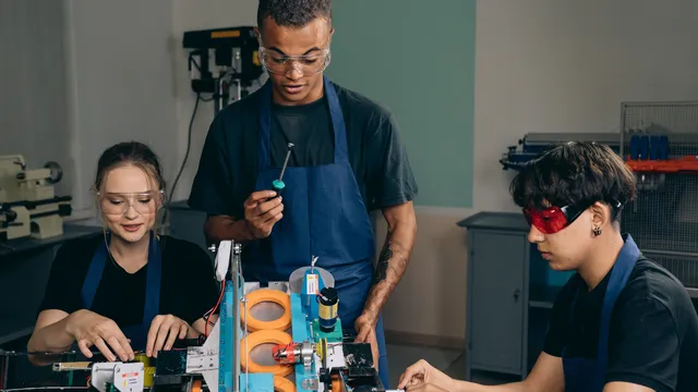 Ein Mitarbeiter in Schutzbrille zeigt zwei Kolleginnen in der Werkstatt einen Handgriff am Gerät