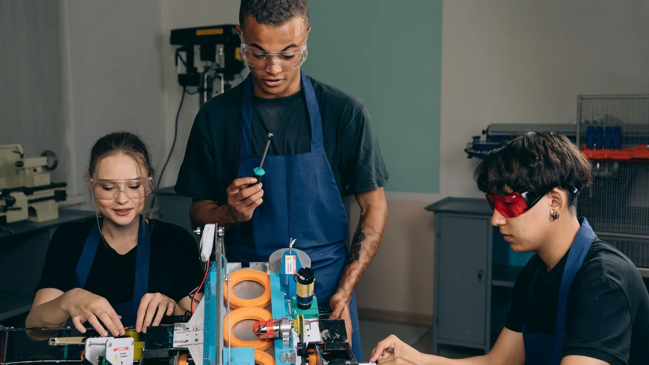 Ein Mitarbeiter in Schutzbrille zeigt zwei Kolleginnen in der Werkstatt einen Handgriff am Gerät